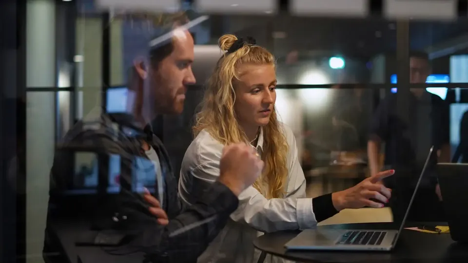 Två kollegor samarbetar vid en laptop i ett modernt kontor och diskuterar något på skärmen.