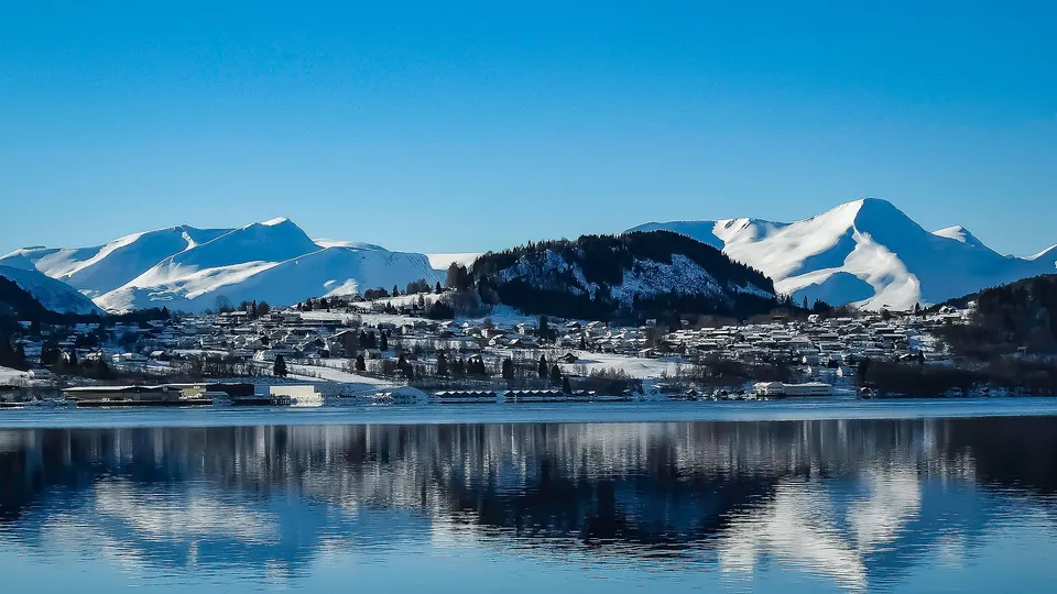 Snøkledde fjell og kystby reflektert i stille fjordvann.