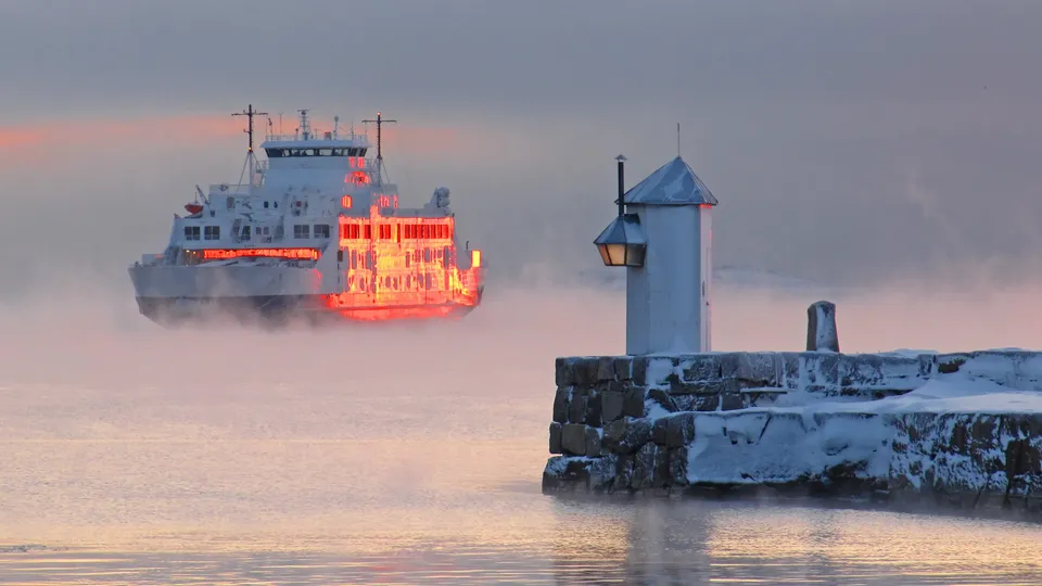 Isbryter med opplyste vinduer i tåkete farvann ved snødekt fyrtårn i skumringen.