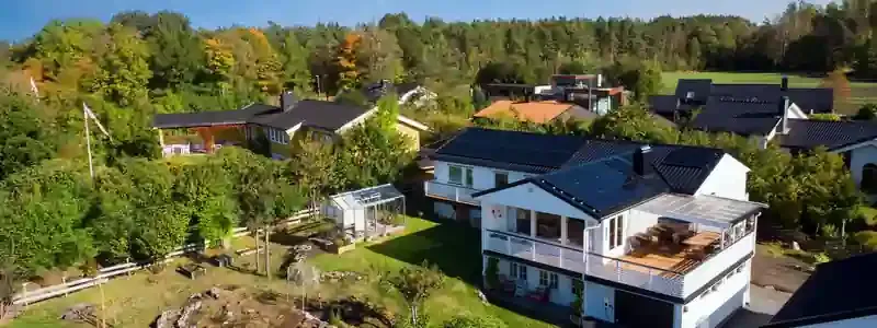 Una casa bianca a due piani con pannelli solari sul tetto scuro, circondata da giardini e alberi.