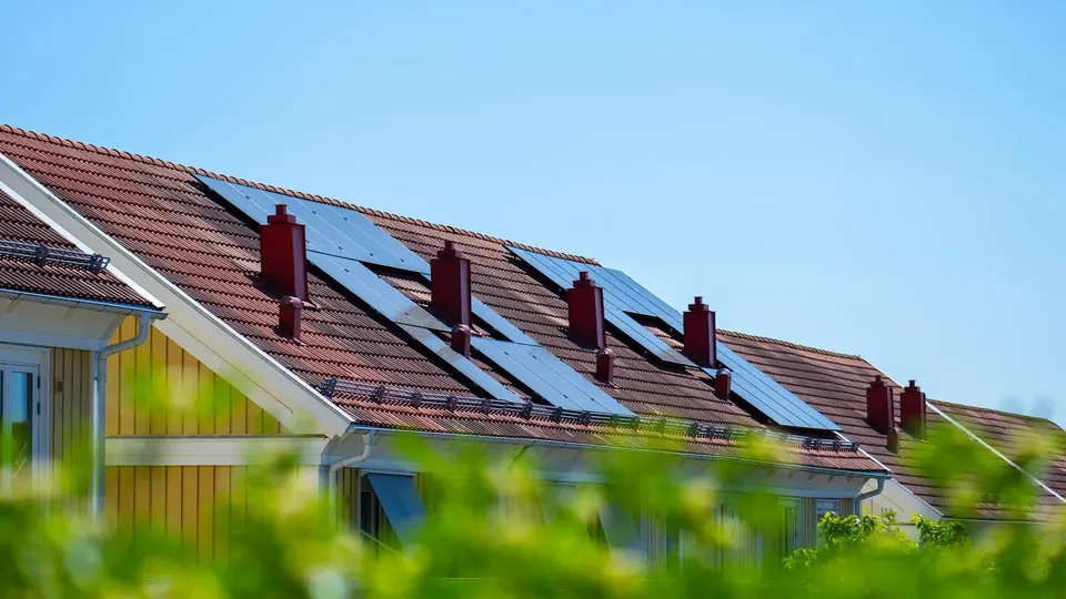 Solens strålar träffar solpanelerna på ett villatak, med somrig grönska i förgrunden.