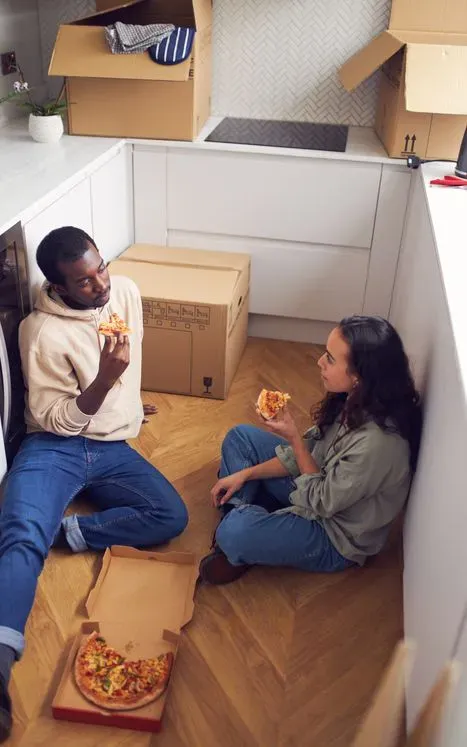Två personer sitter på golvet och äter pizza omgivna av flyttkartonger.