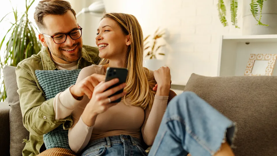 Par smiler mens de ser på smartphone sammen på sofa.