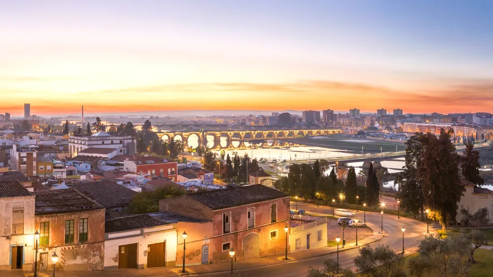 Plaza histórica al atardecer con vistas al puente acueducto romano iluminado y río.