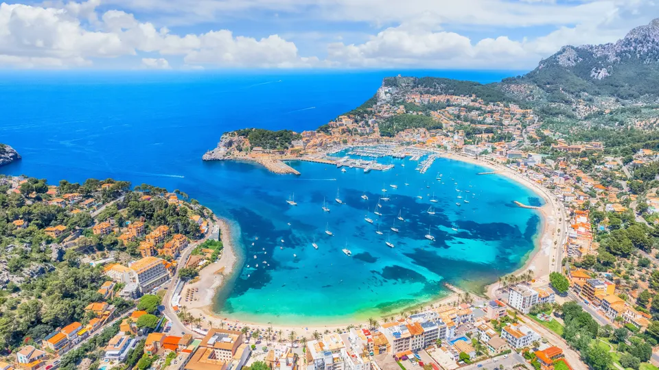 Vista aérea de un pueblo costero mediterráneo con bahía turquesa y veleros amarrados.