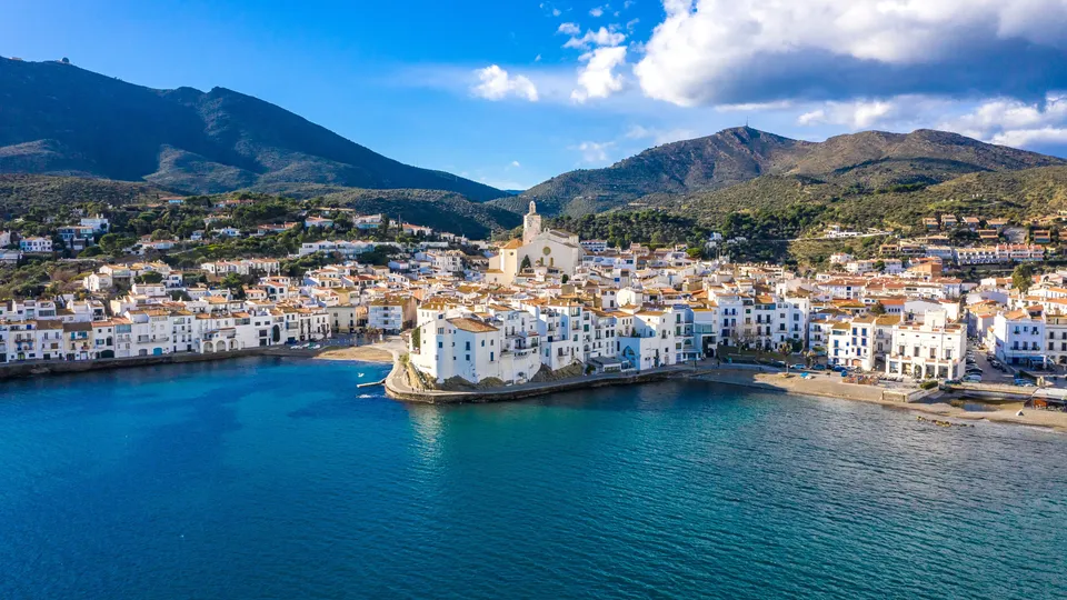 Pueblo costero mediterráneo con edificios blancos y amarillos entre montañas verdes.