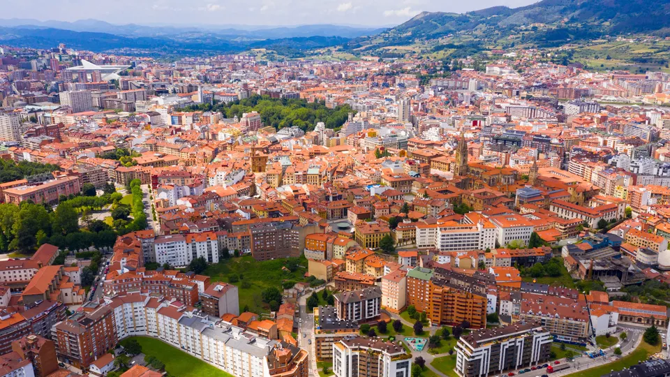 Vista aérea de la ciudad de Oviedo con edificios de tejados naranjas y montañas circundantes.