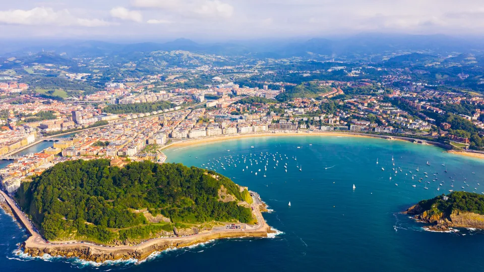 Vista aérea de ciudad costera con playa curva, veleros en bahía turquesa, promontorio boscoso y montañas circundantes.