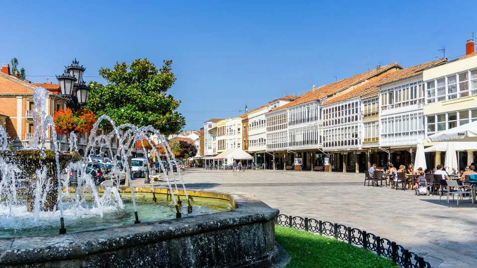 Fuente con agua brotando en una plaza rodeada de edificios históricos y mesas al aire libre.