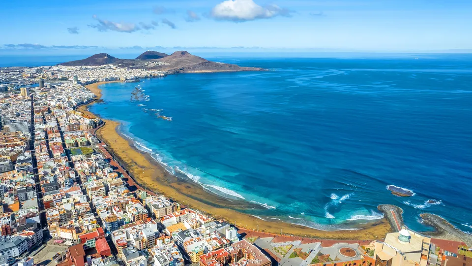 Vista aérea de ciudad costera con playa y montañas volcánicas frente al océano azul.