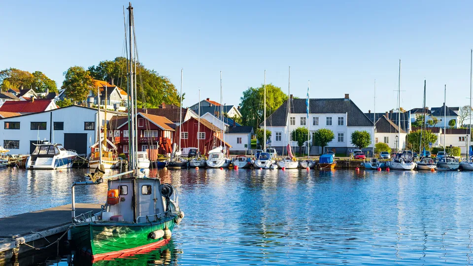 Grønn og rød fiskebåt fortøyd ved marina med seilbåter og hus ved vannkanten.