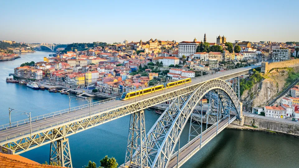 Comboio amarelo atravessando ponte de arco metálico sobre rio azul com cidade colorida na encosta ao fundo.