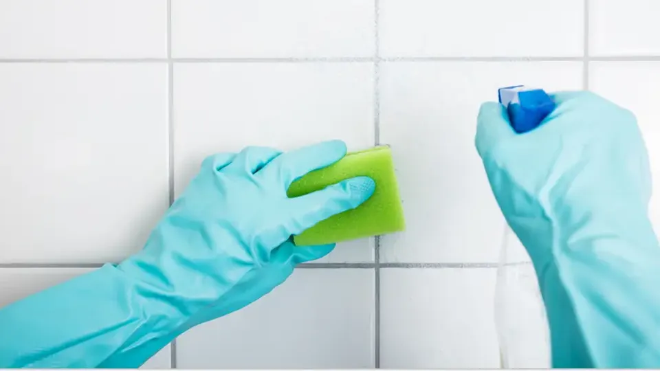 En person med städhandskar rengör vitt kakel med svamp och sprayflaska.