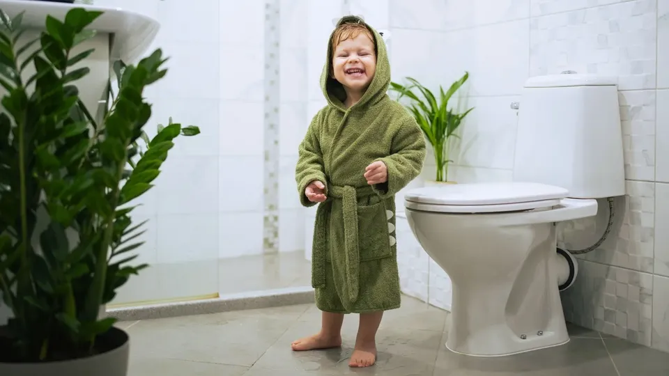 En liten kille i en grön badrock står bredvid toaletten i familjens badrum.