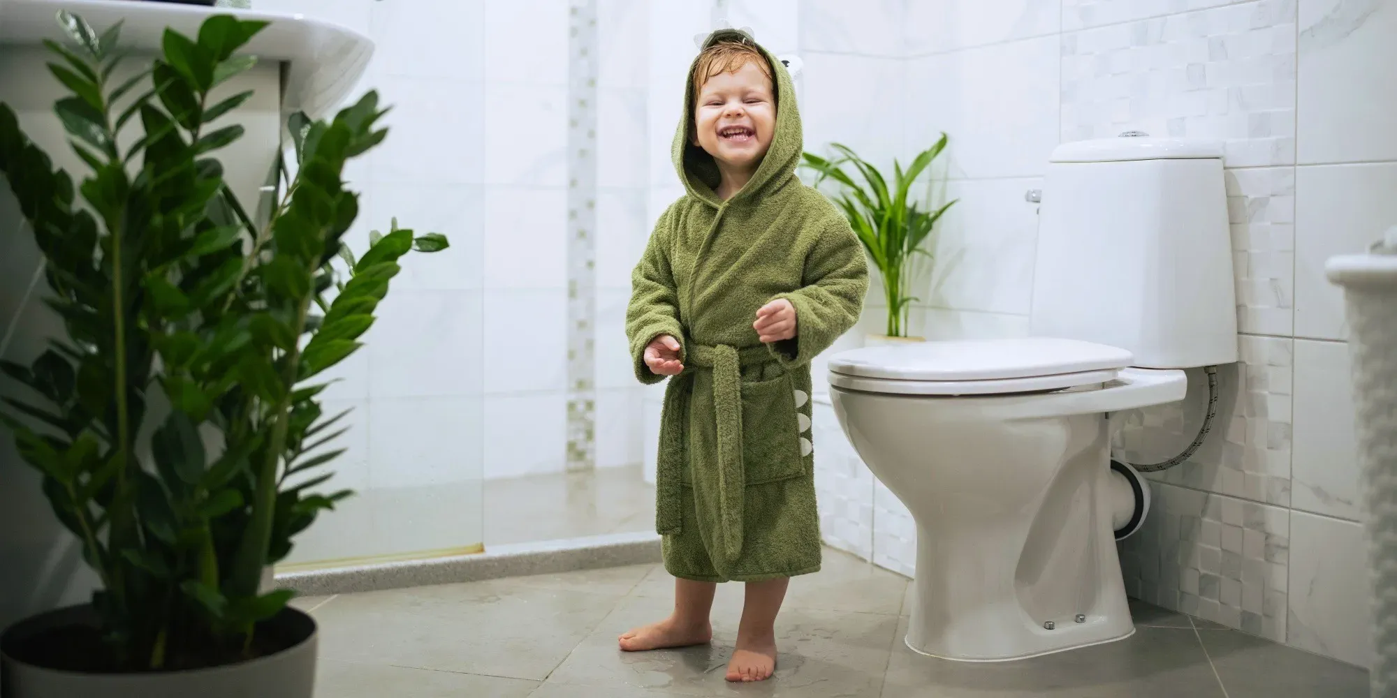 En liten kille i en grön badrock står bredvid toaletten i familjens badrum.