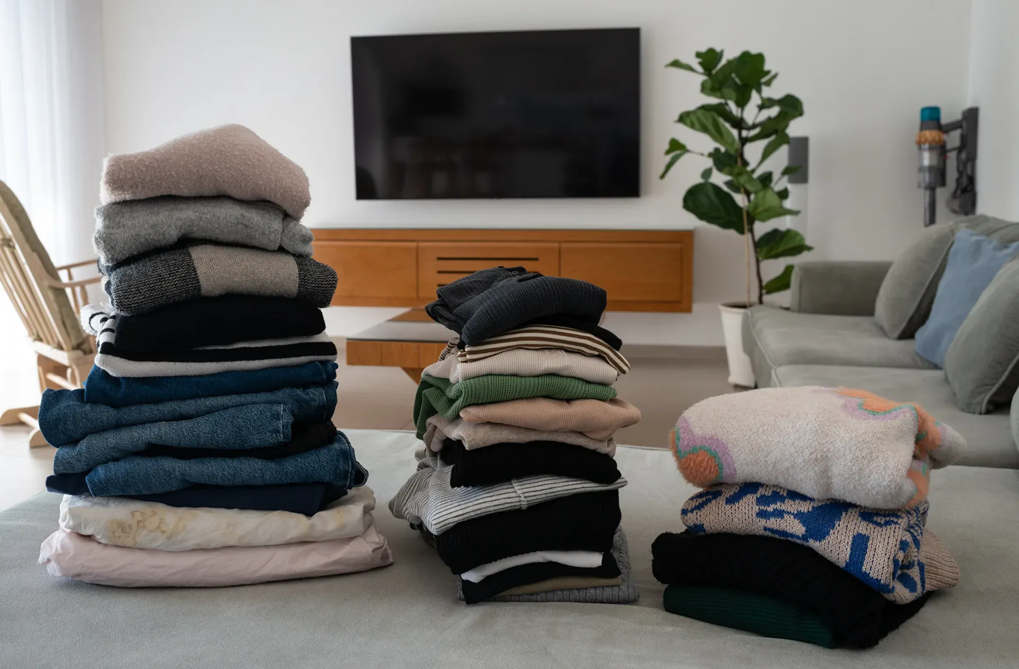 Tre högar med vikta kläder i olika färger ligger på en soffa i ett ljust och stilrent vardagsrum med TV, träbänk och grön växt i bakgrunden.