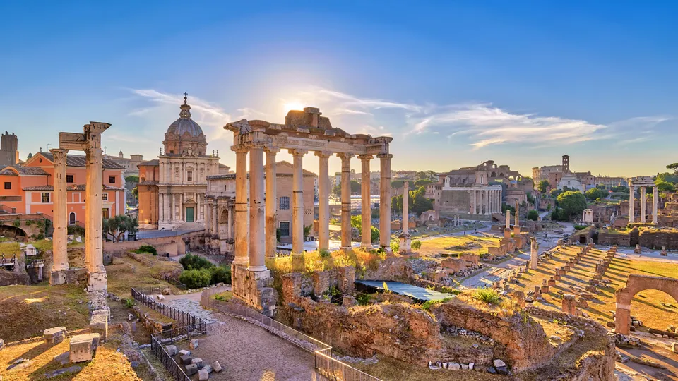 Rovine romane antiche con colonnato e edifici storici sotto cielo blu.