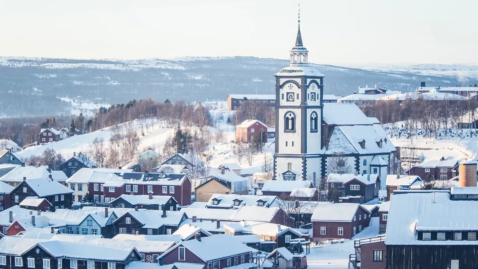Snødekt by med blå og hvit kirketårn som reiser seg over tradisjonelle trehus og skogkledde fjell.