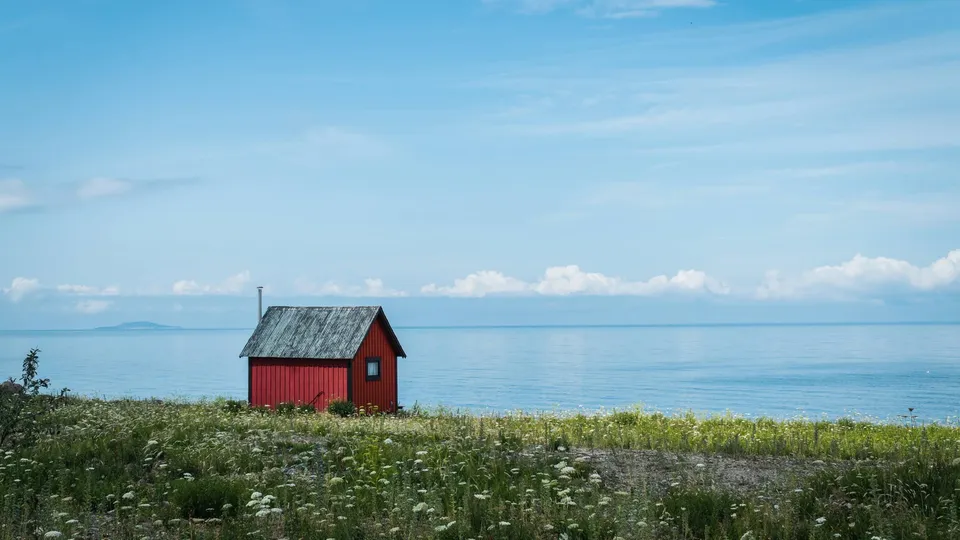 En liten röd stuga vid havet, omgiven av grönska och vilda blommor under en blå himmel.