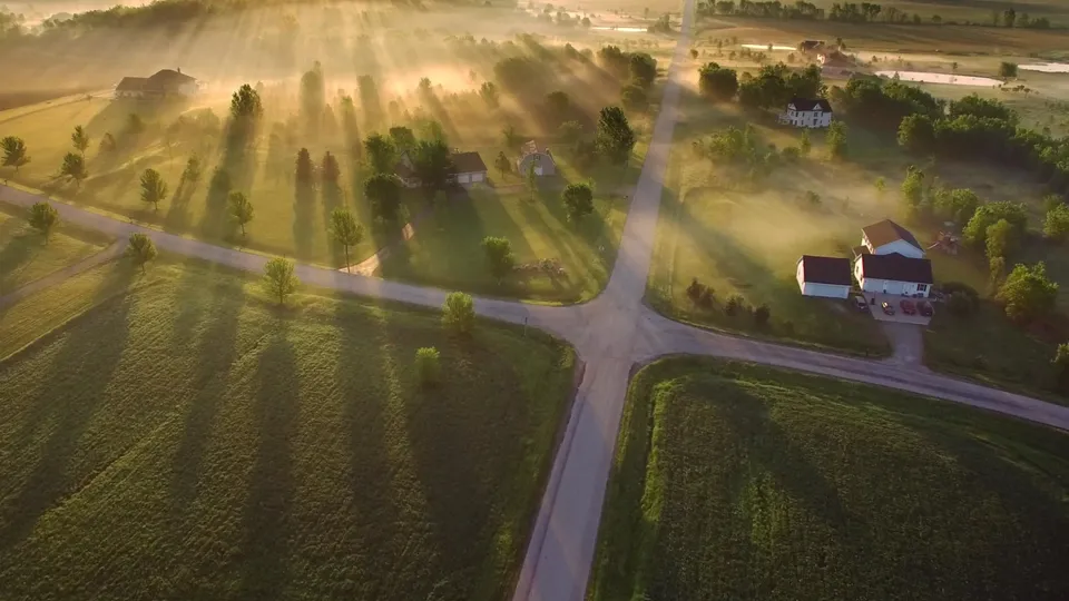 Flygfoto över lantligt jordbruksland och utspridda hus i dimmigt morgonljus.