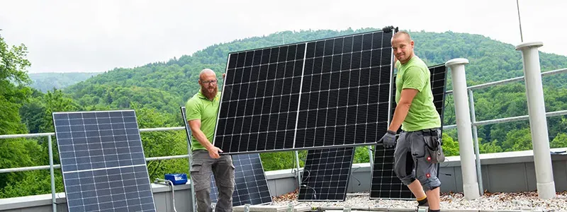 SalzachSonne GmbH – zwei Monteure tragen Photovoltaikmodul auf Flachdach vor grüner Hügellandschaft
