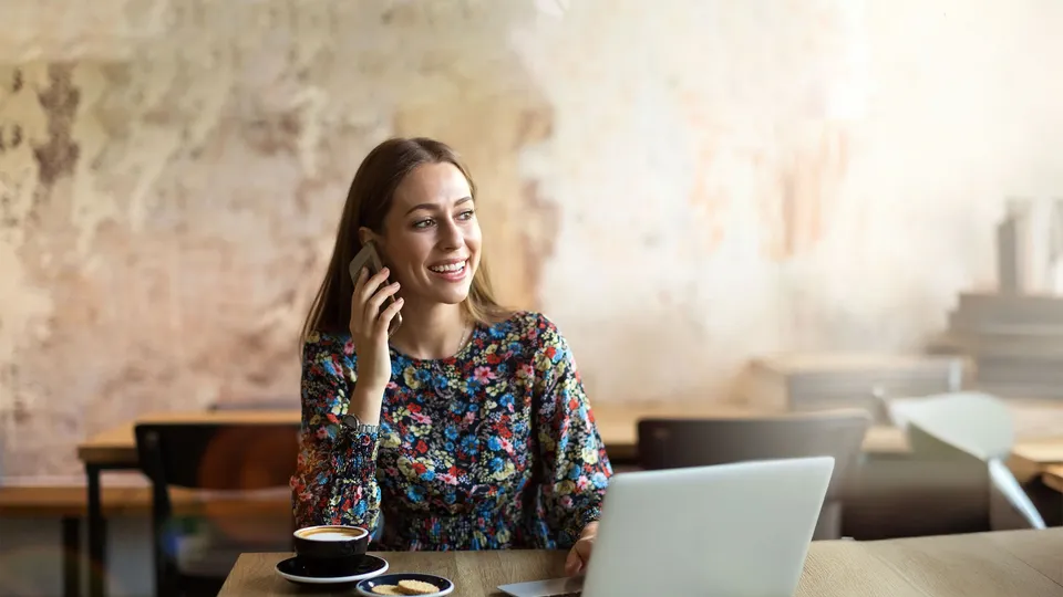 Kvinde taler i telefon ved laptop med kaffekop.