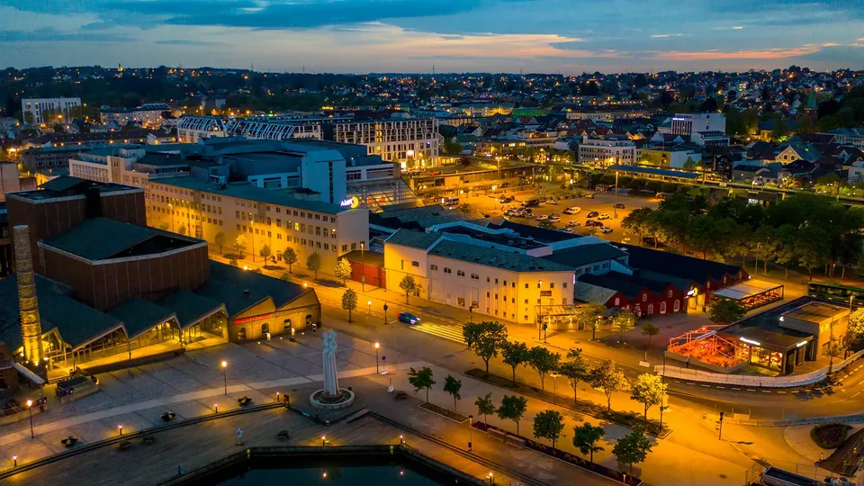 Aerial view of a city center at dusk, with illuminated streets, historic industrial buildings, and a modern shopping mall surrounding a public square and pond.