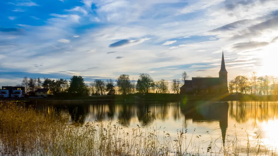 A church with a tall spire silhouetted against a golden sunset, reflected in a calm lake surrounded by reeds and trees, with houses visible on the shore.