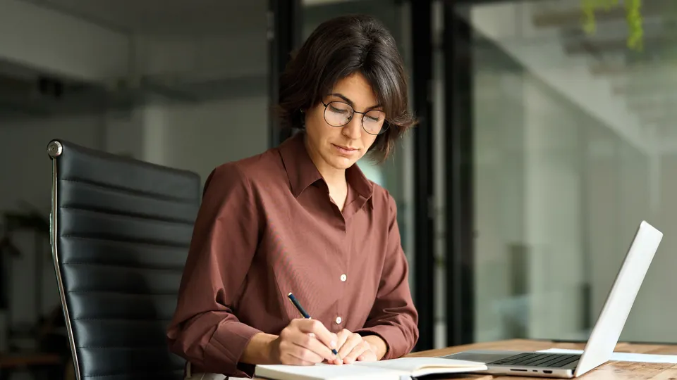 Mujer escribiendo en un cuaderno mientras trabaja en el escritorio con una computadora portátil.