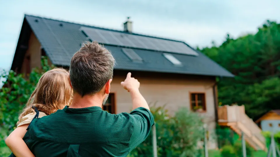 Hombre sosteniendo a un niño mientras señala una casa con paneles solares.