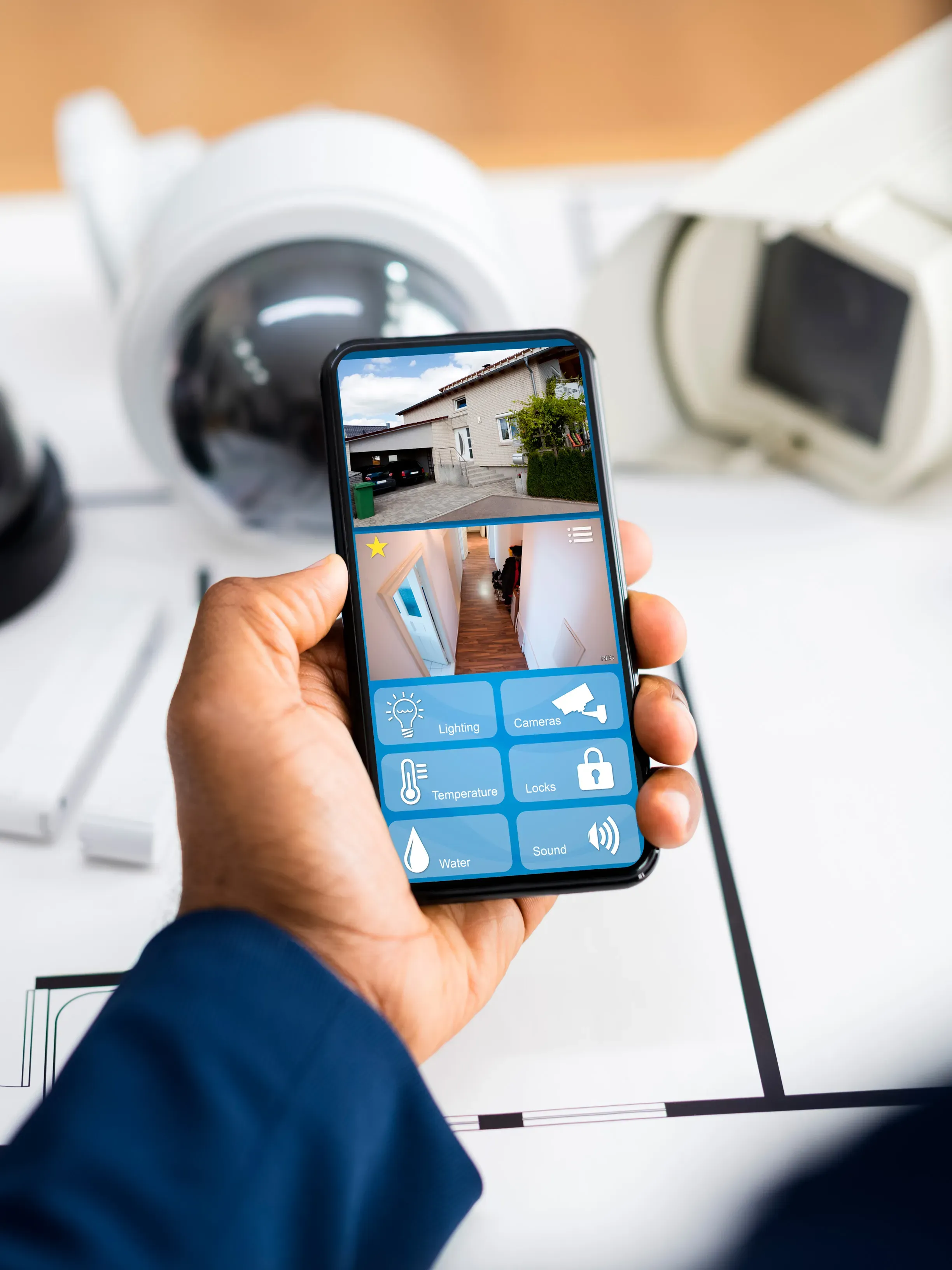 Persona sosteniendo smartphone mostrando aplicación de control del hogar inteligente con cámaras de seguridad y automati.