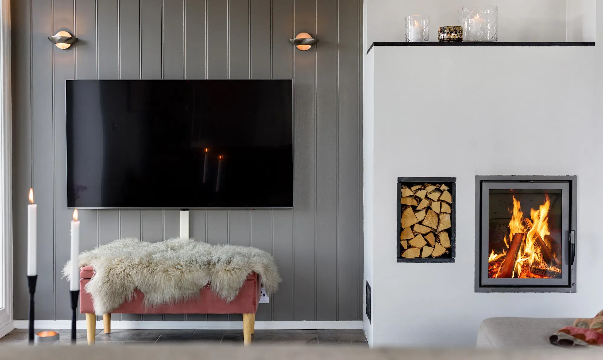 Veggmontert TV over en rosa benk med saueskinn ved siden av en peis med brennende ved.