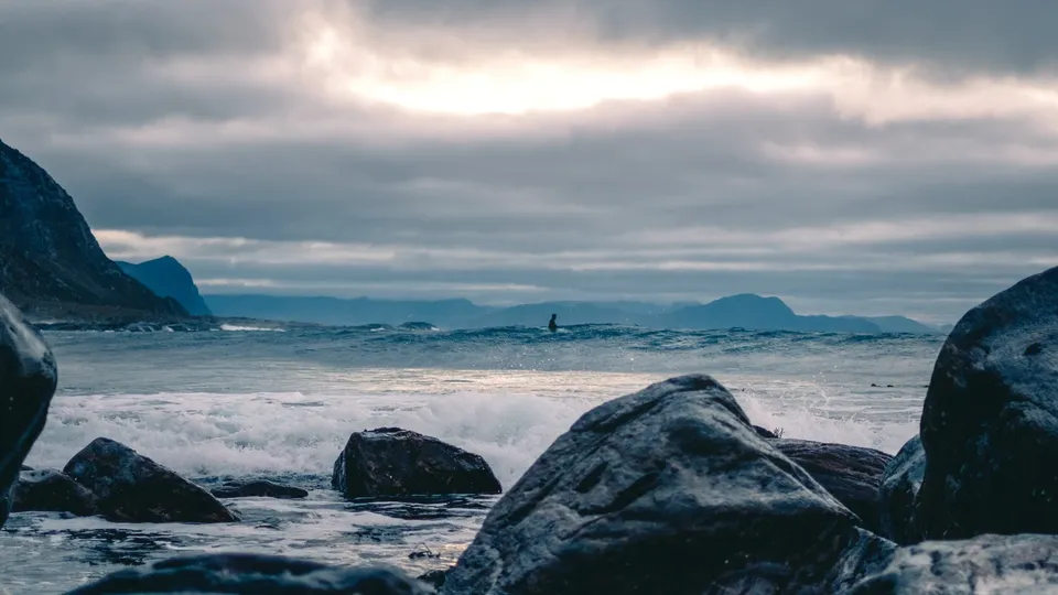 Surfer i havbølger med steinete kystlinje og fjell i bakgrunnen - sjøvarme.