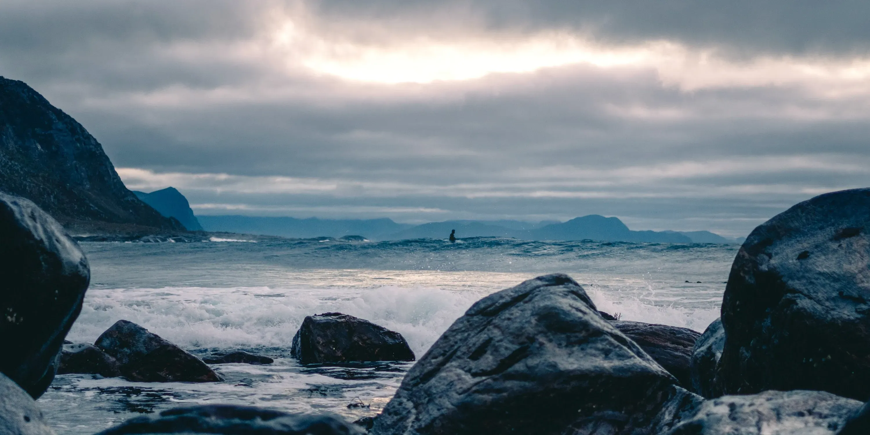 Surfer i havbølger med steinete kystlinje og fjell i bakgrunnen - sjøvarme.