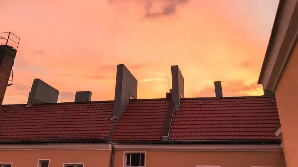 Tak med röda tegelpannor och skorstenar mot orange solnedgångshimmel.