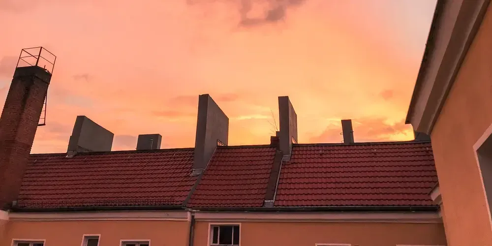Tak med röda tegelpannor och skorstenar mot orange solnedgångshimmel.