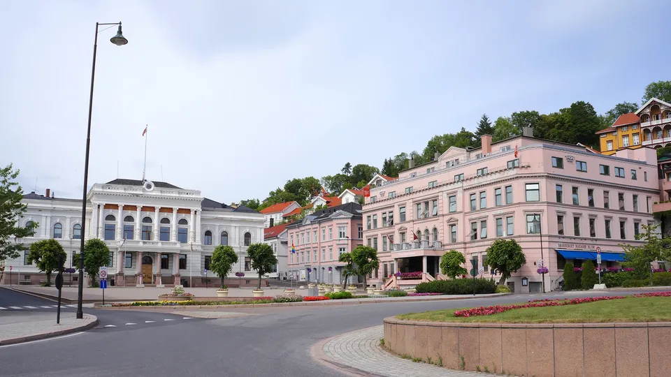 Torget med hvitt rådhus med søyler, rosa hotellbygning og fargede hus på skråningen.