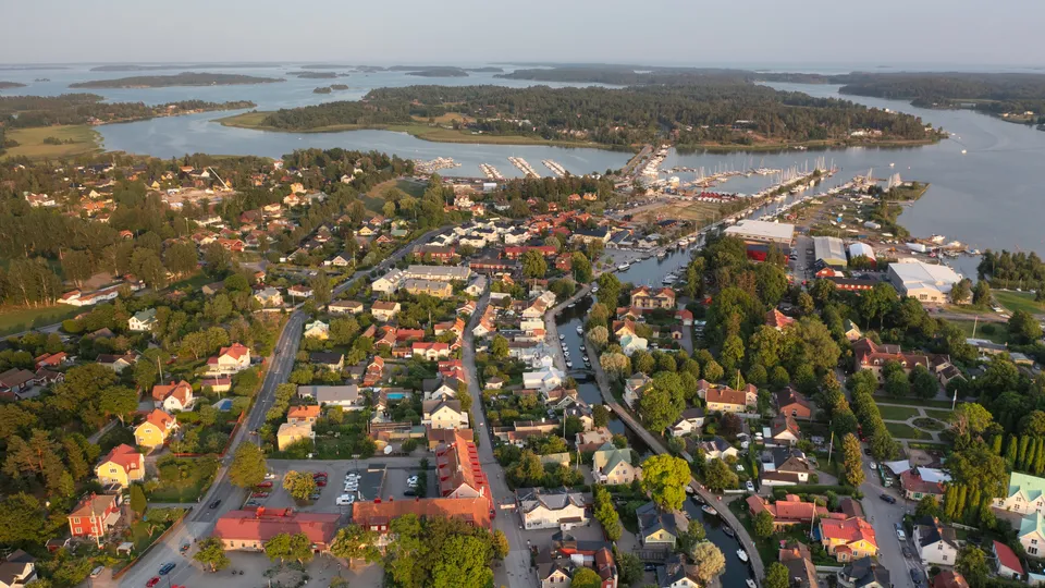Flygfoto över Trosa med rödtakade hus, skogsområden och en båthamn med segelfartyg.