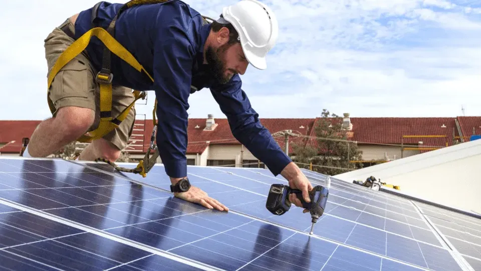 Worker installing solar panels on rooftop using power drill.