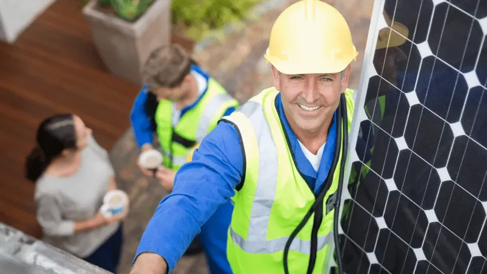 Solar panel installer in safety gear smiling at camera next to solar panel.