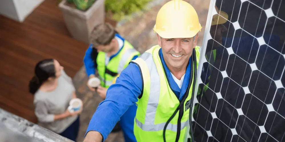 Solar panel installer in safety gear smiling at camera next to solar panel.