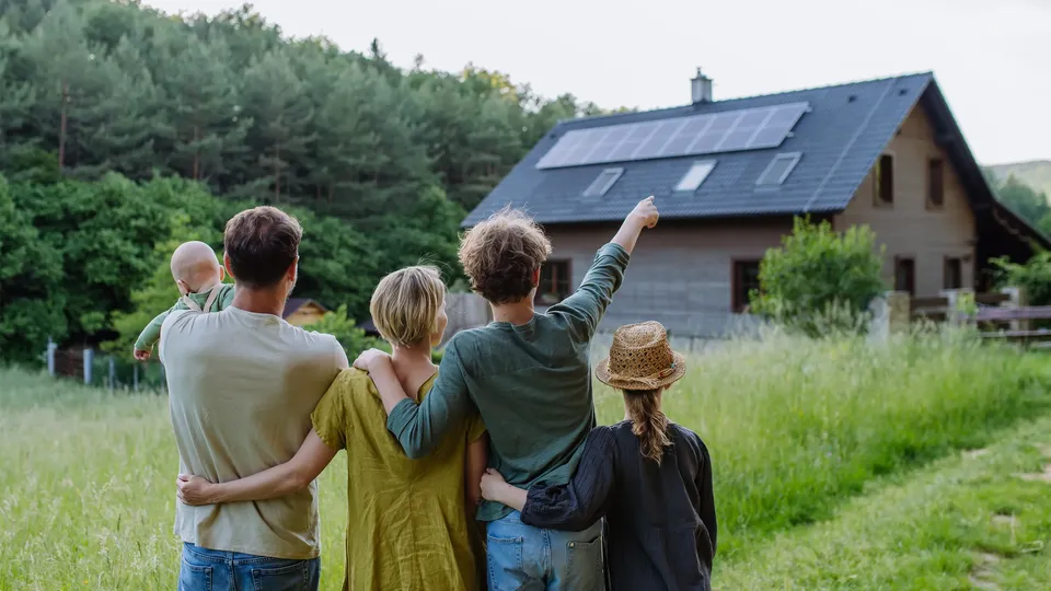 Fünfköpfige Familie steht auf Wiese und blickt auf Haus mit Solaranlage auf dem Dach.