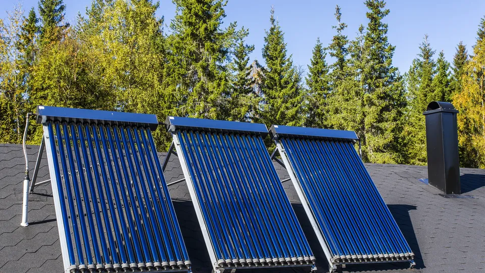 Solarthermieanlage mit drei Vakuumröhrenkollektoren auf einem Hausdach vor einem Hintergrund aus Nadelbäumen und blauem Himmel.