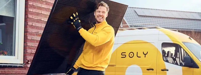"Soly – lächelnder Monteur trägt Photovoltaikmodul vor gelbem Firmenfahrzeug