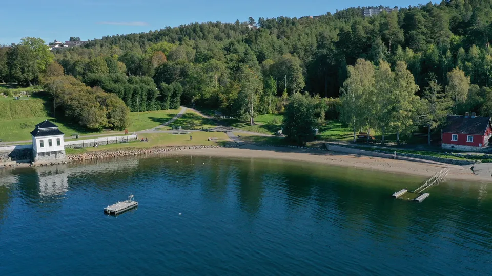 Malerisk lakebygd med sandstrand, hvitt båthus, rød hytte og skogkledd fjellside.