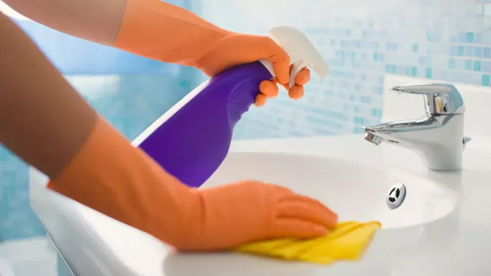 Person med orange städhandskar sprayar rengöringsmedel i ett handfat.