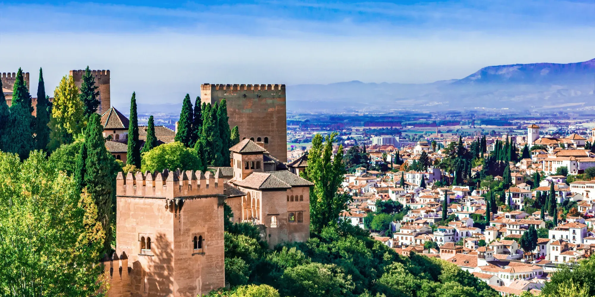 Torres de fortaleza medieval dominan el paisaje urbano de Granada con cipreses y montañas al fondo.