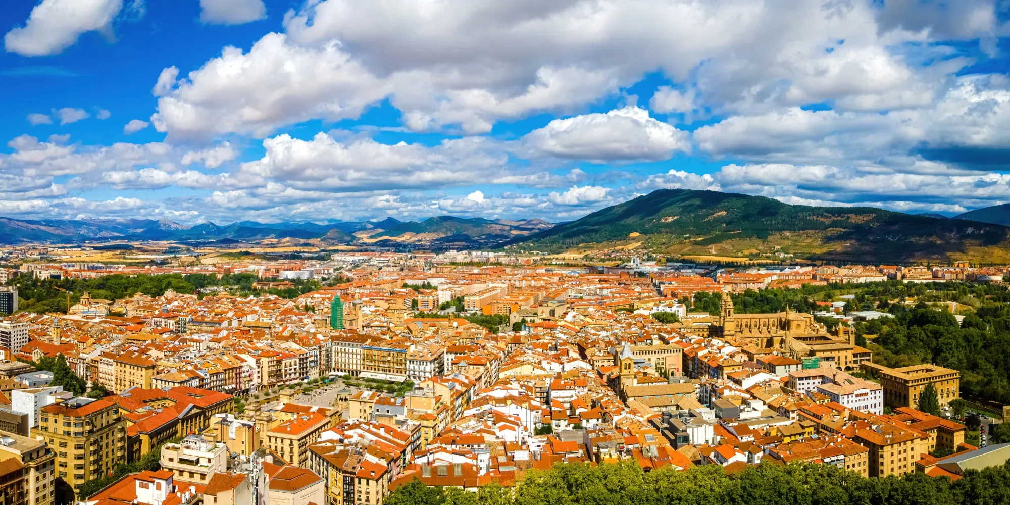 Vista aérea de una ciudad con tejados de tejas naranjas y una montaña verde al fondo.