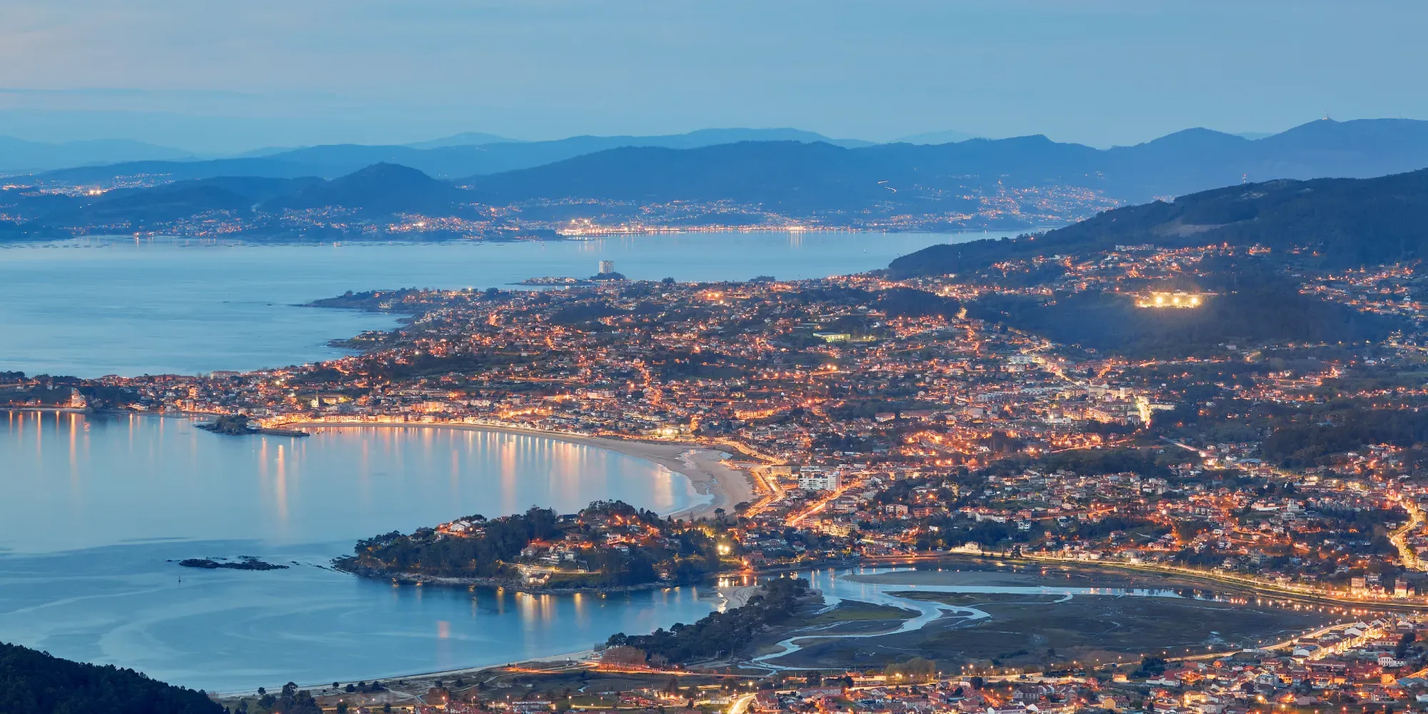 Ciudad costera al atardecer con edificios iluminados rodeando una bahía y montañas distantes.