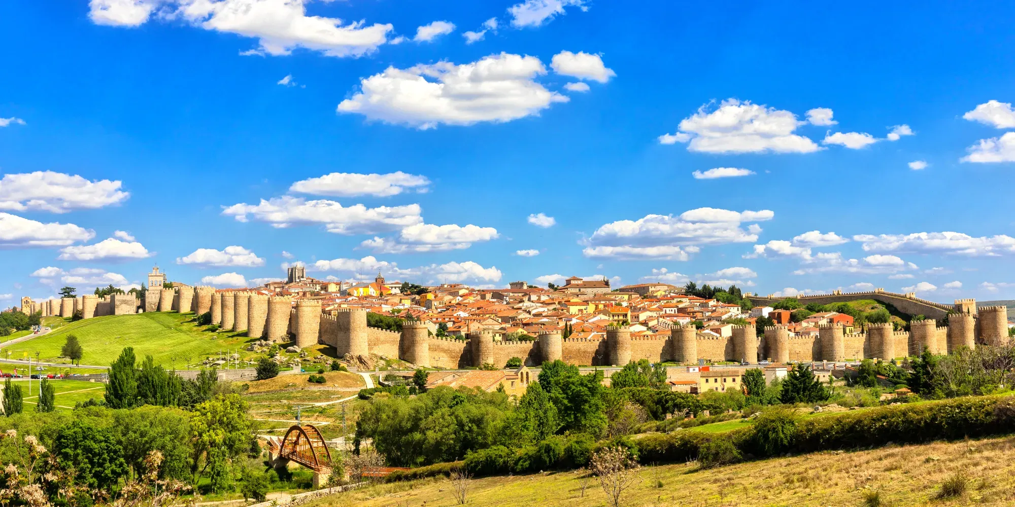 Ciudad medieval amurallada con fortificaciones de piedra y tejados de terracota bajo cielo azul.